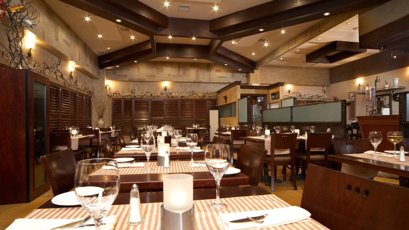 Elegant restaurant interior with exposed dark wood beam ceiling, natural travertine stone accent walls, white tablecloths on dark wood tables, brown leather upholstered chairs, warm ambient lighting, and contemporary design in Bexleyheath