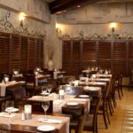 Thumbnail of http://Restaurant%20side%20dining%20area%20with%20textured%20beige%20stone%20walls,%20dark%20wood%20plantation%20shutters,%20decorative%20wrought%20iron%20vine%20motif%20along%20the%20wall,%20dark%20wood%20tables%20with%20striped%20runners%20and%20candles,%20and%20warm%20ceiling%20spotlights%20in%20Bexleyheath