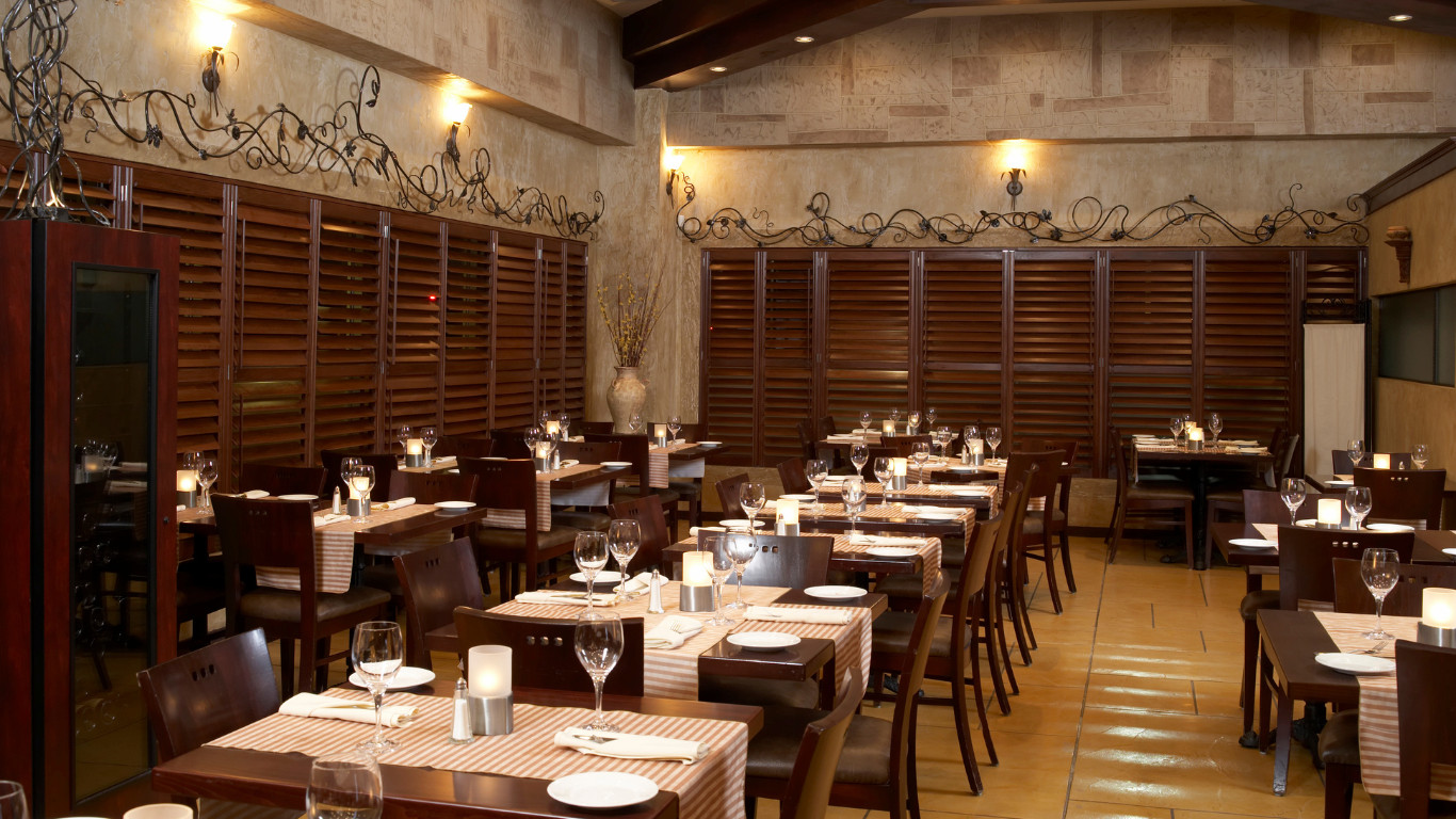 Restaurant side dining area with textured beige stone walls, dark wood plantation shutters, decorative wrought iron vine motif along the wall, dark wood tables with striped runners and candles, and warm ceiling spotlights in Bexleyheath