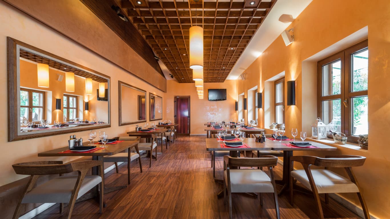 Elegant restaurant interior with warm neutral design, natural wood dining tables and chairs, golden amber walls, soft ambient lighting, large windows with natural light, comfortable seating, earthy colour scheme, and modern rustic aesthetic in Dulwich