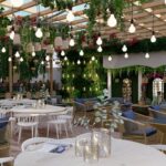 Thumbnail of http://High-end%20restaurant%20interior%20with%20cascading%20hanging%20plants%20and%20vines%20from%20ceiling,%20vintage%20Edison%20bulb%20string%20lights,%20natural%20wood%20built-in%20banquette%20seating%20with%20blue%20cushions,%20white%20modern%20dining%20tables%20and%20chairs,%20potted%20plants,%20arched%20windows%20with%20greenery%20views,%20and%20light-filled%20airy%20atmosphere%20in%20Notting%20Hill