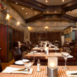 Thumbnail of http://Natural%20toned%20restaurant%20interior%20featuring%20dark%20wood%20beam%20ceiling%20with%20spotlights,%20textured%20beige%20stone%20walls,%20dark%20wood%20tables%20with%20beige%20and%20white%20striped%20runners,%20brown%20leather%20chairs,%20candlelit%20centerpieces,%20and%20warm%20ambient%20lighting%20in%20Bexleyheath