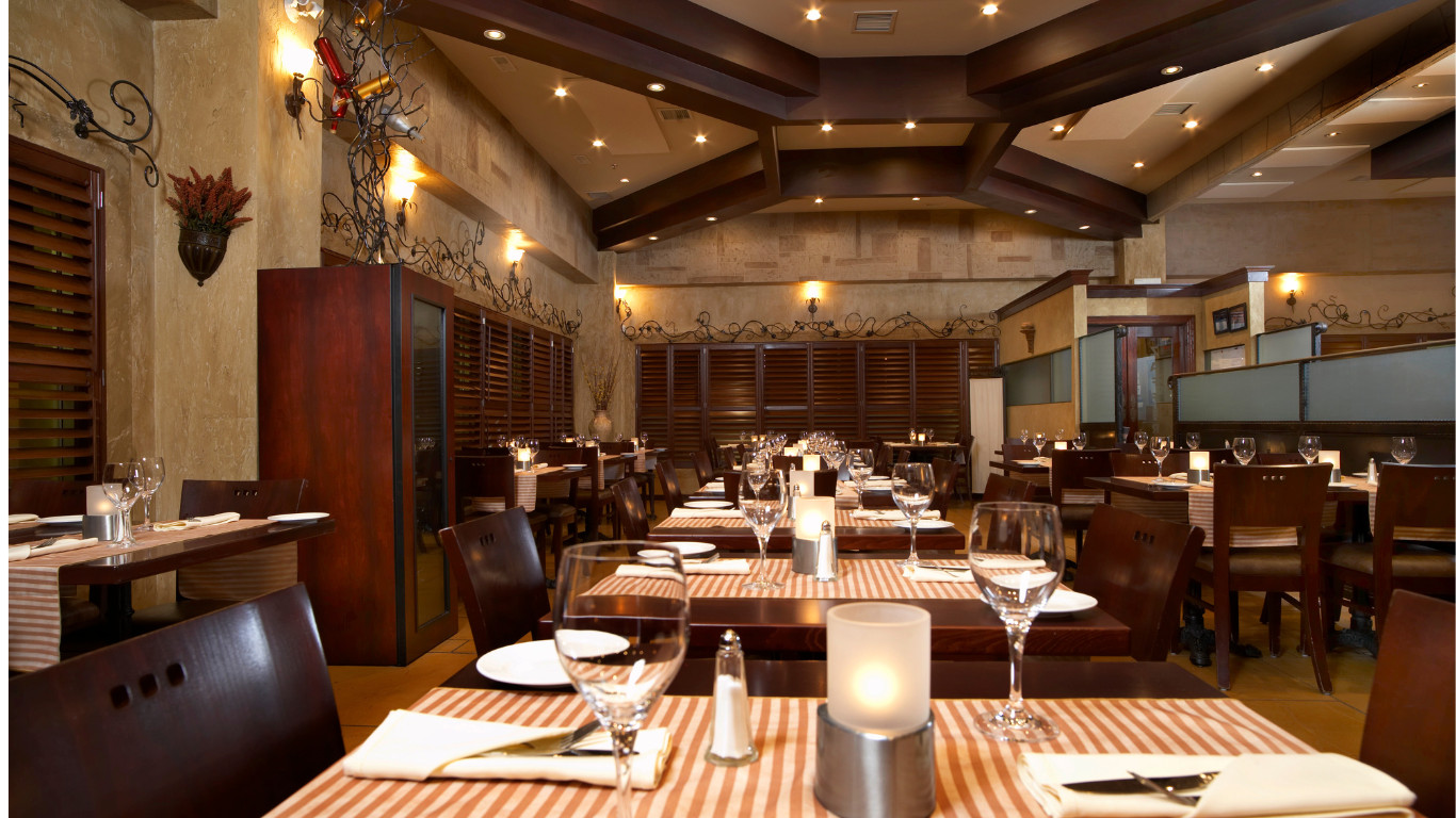 Natural toned restaurant interior featuring dark wood beam ceiling with spotlights, textured beige stone walls, dark wood tables with beige and white striped runners, brown leather chairs, candlelit centerpieces, and warm ambient lighting in Bexleyheath