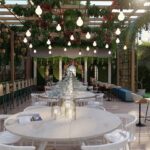 Thumbnail of http://Elegant%20restaurant%20corridor%20with%20hanging%20plants%20and%20vines%20overhead,%20vintage%20Edison%20bulb%20string%20lights,%20natural%20wood%20banquette%20seating%20with%20green%20cushions%20on%20both%20sides,%20long%20white%20circular%20communal%20dining%20table%20in%20center,%20potted%20plants,%20large%20windows%20with%20garden%20views,%20and%20light-filled%20airy%20atmosphere%20in%20Notting%20Hill