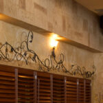 Thumbnail of http://Restaurant%20architectural%20detail%20featuring%20travertine%20stone%20wall,%20decorative%20wrought%20iron%20scrollwork%20mounted%20above%20horizontal%20dark%20wood%20slat%20panels,%20wall%20sconces%20with%20amber%20shades,%20and%20warm%20ambient%20lighting%20in%20Bexleyheath