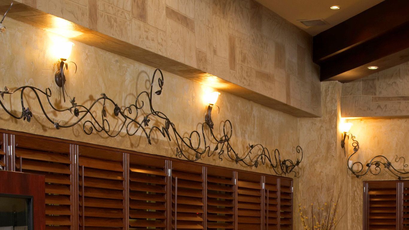 Restaurant architectural detail featuring travertine stone wall, decorative wrought iron scrollwork mounted above horizontal dark wood slat panels, wall sconces with amber shades, and warm ambient lighting in Bexleyheath