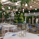 Thumbnail of http://Wide-angle%20view%20of%20high-end%20restaurant%20interior%20with%20cascading%20hanging%20plants%20and%20vines%20from%20ceiling,%20vintage%20Edison%20bulb%20string%20lights,%20natural%20wood%20banquette%20seating%20with%20blue%20and%20green%20cushions%20along%20walls,%20white%20modern%20dining%20tables%20in%20foreground,%20glass%20partition%20with%20greenery,%20arched%20windows,%20and%20light-filled%20spacious%20atmosphere%20in%20Notting%20Hill
